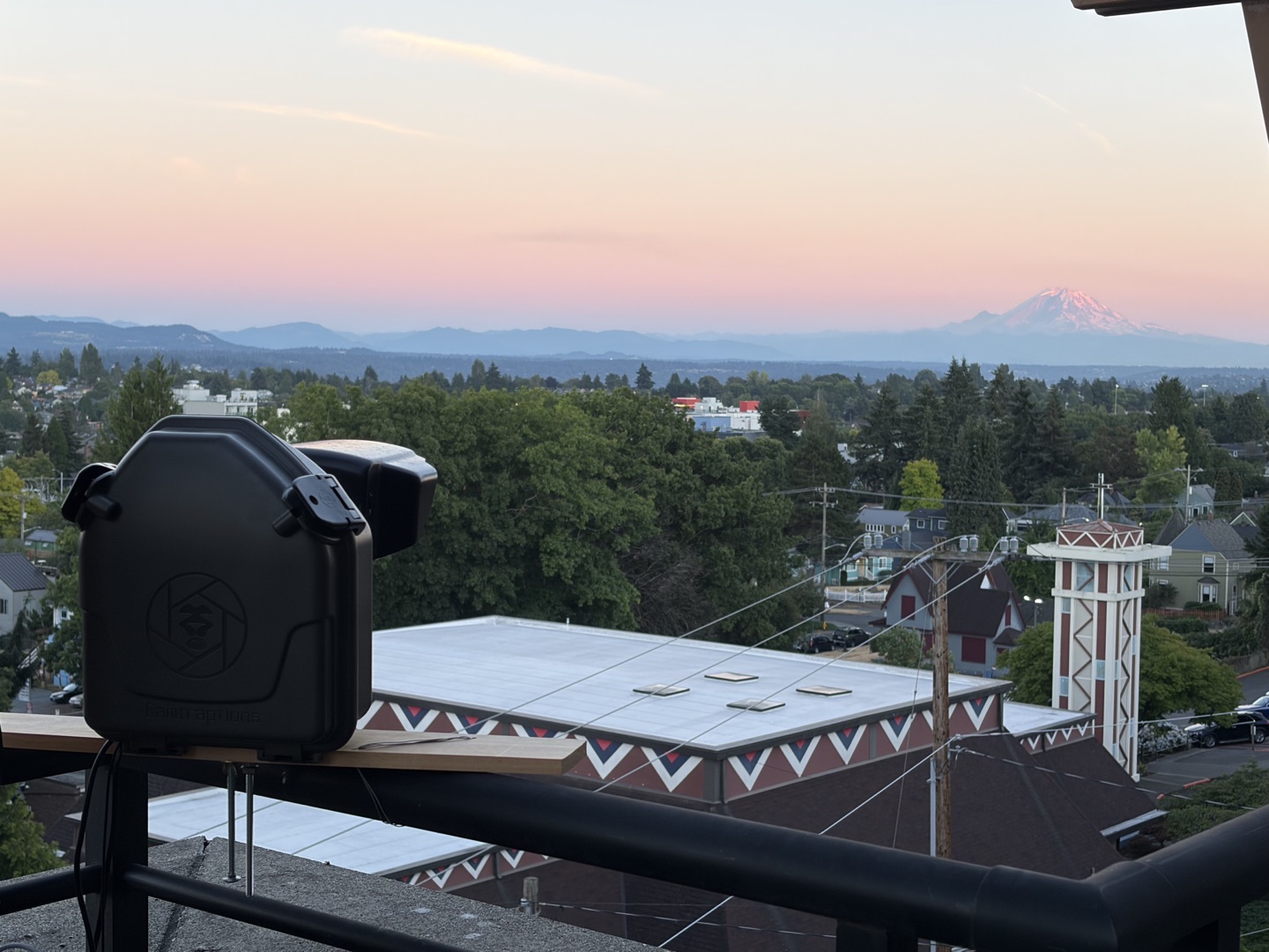 The camera in its weatherproof housing on a balcony rail at sunset, with Mount Rainier lit pink on the horizon beyond the neighborhood